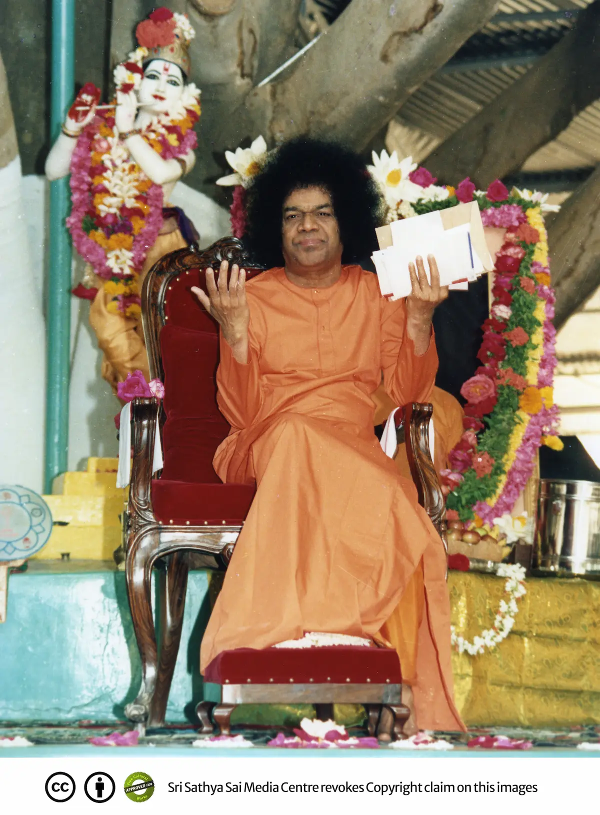 Photograph of Sathya Sai Baba seated on an ornate chair at Brindavan, dressed in an orange robe and holding papers, surrounded by decorative elements